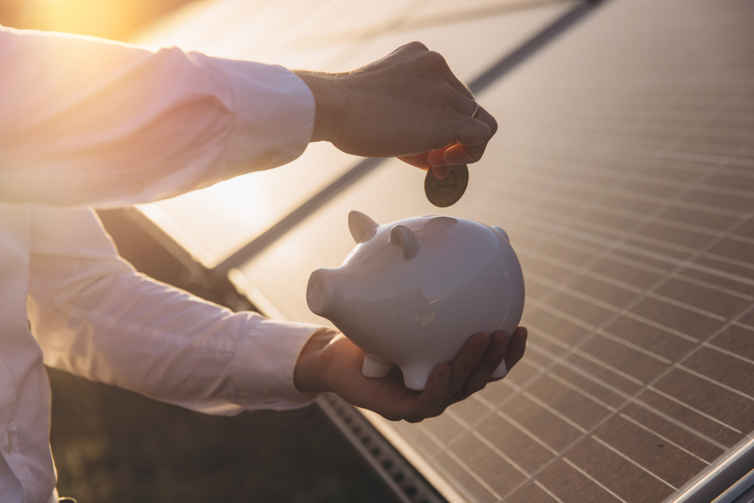 A person places a coin into a piggy bank against the backdrop of solar panels at sunset, symbolizing financial savings and renewable energy investments.