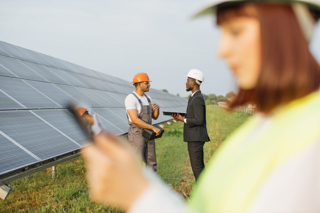 African man in suit and indian technician in uniform controlling work of solar cells while standing together on field. Blur foreground of woman in helmet working on digital tablet.